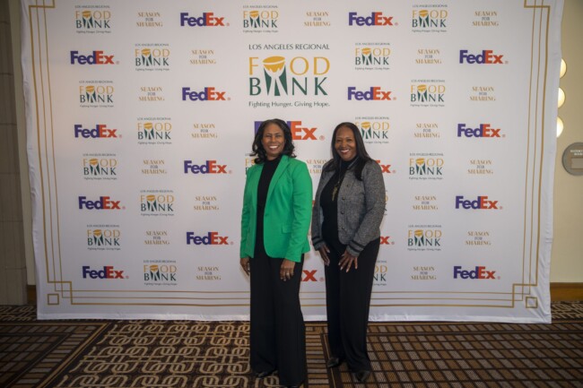 Two women smiling in front of a backdrop featuring branding for the Los Angeles Regional Food Bank and FedEx. They are standing on a patterned carpet, and one is wearing a green blazer while the other is in a dark suit.