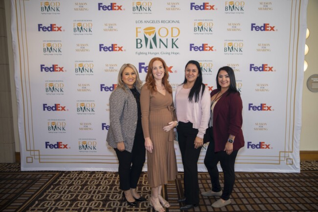 Four women stand together smiling in front of a backdrop with logos for the Los Angeles Regional Food Bank and FedEx. They are on a patterned carpet and are dressed in business casual attire.