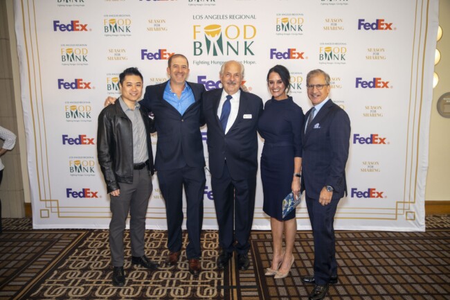 A group of five people stands smiling in front of a backdrop with Los Angeles Regional Food Bank and FedEx logos. They are dressed in formal attire, with four men and one woman posing together.