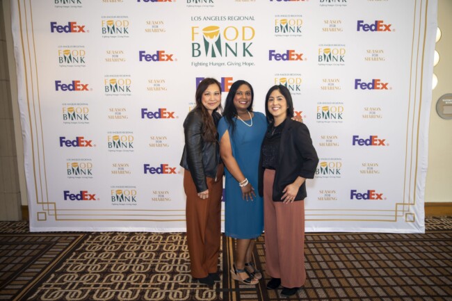 Three women pose together smiling in front of a backdrop featuring logos for the Los Angeles Regional Food Bank and FedEx. The backdrop displays the Food Bank's slogan, "Fighting Hunger, Giving Hope.
