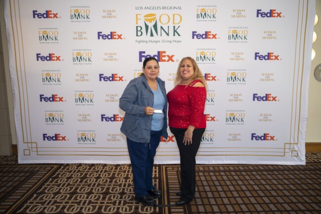 Two women stand in front of a backdrop with logos for the Los Angeles Regional Food Bank and FedEx. One is wearing a red top, and the other is in a blue jacket holding a piece of paper. The event appears formal and supportive.