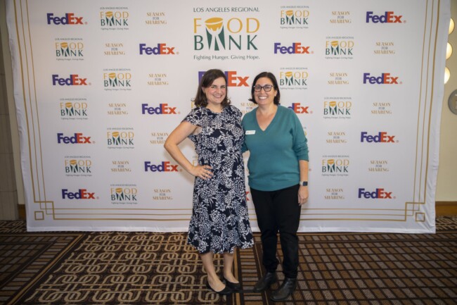 Two women smiling and standing in front of a backdrop with logos, including "Los Angeles Regional Food Bank" and "FedEx." They are in a room with checkered carpeting. One wears a patterned dress, and the other wears a teal top.