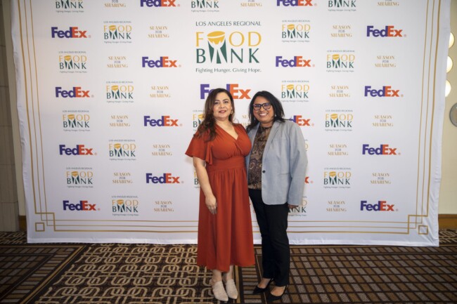 Two women smiling and posing in front of a backdrop with logos for the Los Angeles Regional Food Bank, FedEx, and Season of Sharing. The woman on the left wears a red dress, and the woman on the right wears a gray blazer and glasses.