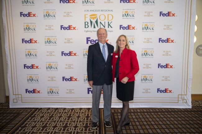 A man in a suit and a woman in a red jacket stand smiling in front of a backdrop featuring logos for the Los Angeles Regional Food Bank and FedEx.