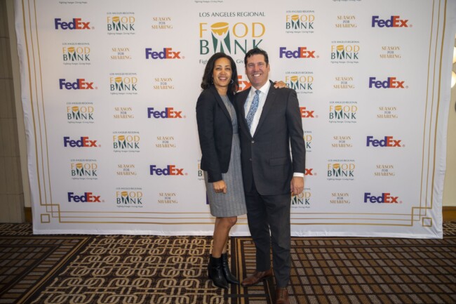 Two people stand smiling in front of a backdrop with logos for the Los Angeles Regional Food Bank, FedEx, and other sponsors. The woman is wearing a black jacket and gray dress, and the man is in a dark suit and tie.