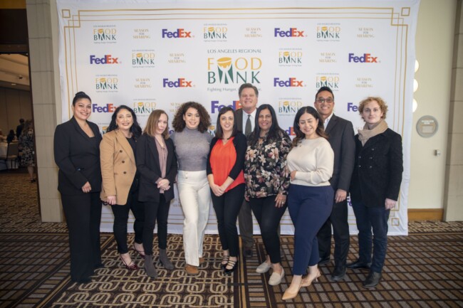 A diverse group of ten people stand smiling in front of a backdrop featuring logos for the Los Angeles Regional Food Bank and FedEx. They are dressed in business casual attire and positioned in a hotel event space.