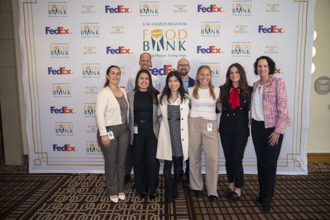 A group of seven people smiling and standing in front of a Los Angeles Regional Food Bank backdrop with FedEx logos. They're dressed in business casual attire, and the carpet has a geometric pattern.