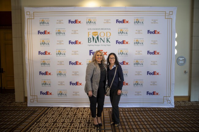 Two people stand smiling in front of a backdrop featuring logos for the Los Angeles Regional Food Bank, FedEx, and other sponsors. The setting appears to be indoors with a patterned carpet underfoot.