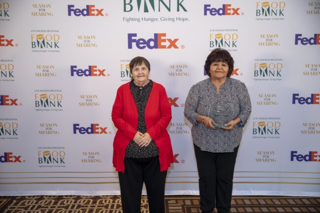 Two women stand side by side at an event. One wears a red cardigan, and the other a patterned blouse. The backdrop features logos for a food bank, FedEx, and "Season for Sharing." The floor has a patterned carpet.