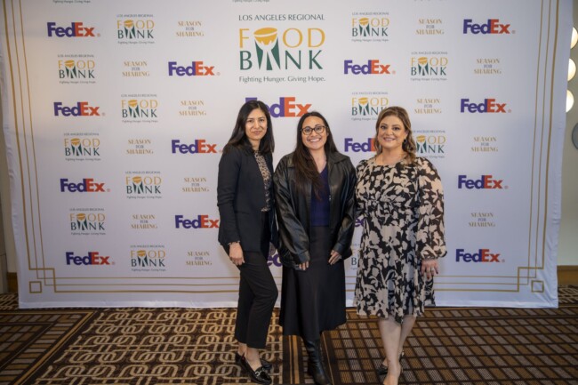 Three women stand in front of a backdrop with logos for FedEx and Los Angeles Regional Food Bank. They are smiling and dressed in professional attire.