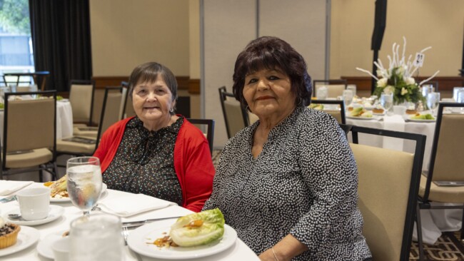 Two women sit at a round dining table set with plates of salads, glasses of water, and centerpiece decorations. They are smiling and casually dressed, with one wearing a red cardigan. The room has several tables and a neutral color scheme.