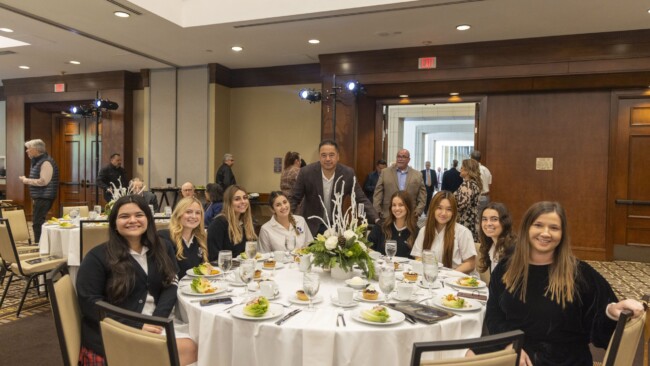 A group of people sits at a round table set for a meal in a banquet hall. They are smiling and dressed in formal attire. A centerpiece with flowers decorates the table. Other guests are visible in the background.