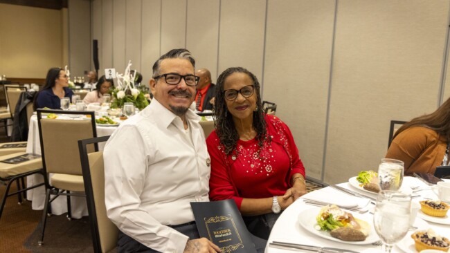 A man in a white shirt and a woman in a red sweater sit together at a banquet table. They are smiling and holding a black booklet. The table is set with glasses, plates, and cutlery, with others dining in the background.
