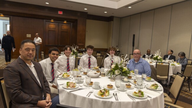 A group of people seated at round tables during a formal event. The tables are set with plates of salad, glasses, and floral centerpieces. Attendees are dressed in formal attire, with several wearing ties. The room has wooden paneling and carpeted floors.