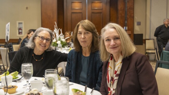 Three women seated at a dining table with plates of food and glasses of water. They are smiling and posing for the photo, with a wooden paneled wall in the background.