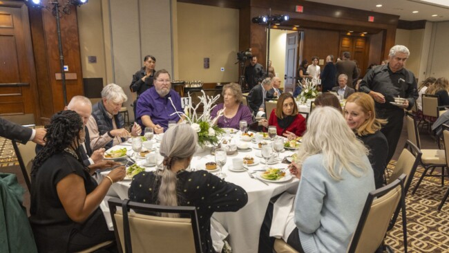 A group of people seated around a round table at a formal event, dining and conversing. The table is elegantly set with plates, glasses, and a flower centerpiece. Other guests and servers are seen in the background.