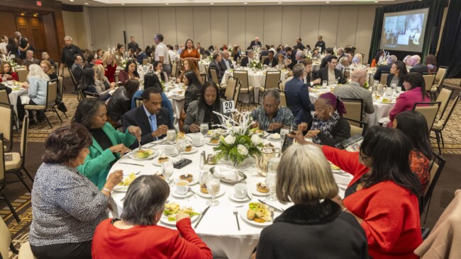 A diverse group of people seated around tables at a formal event, enjoying a meal together. The room is filled with round tables, and a presentation is visible on a screen in the background. Floral centerpieces and place settings adorn each table.