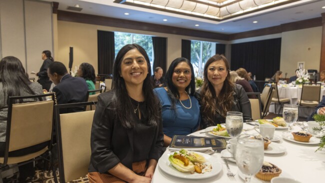 Three women are seated at a dining table, smiling at the camera. Each has a plate of food before them, with other attendees visible in the background in a banquet setting, featuring large windows and elegant decor.
