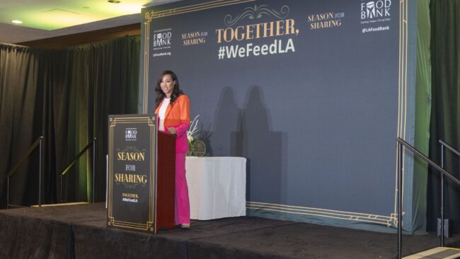 A person stands at a podium on a stage adorned with event branding, including "Together, #WeFeedLA," and "Season for Sharing." They are giving a speech, with a decorative backdrop and Food Bank logos visible.