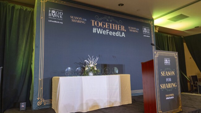 A stage is set for an event with a podium on the right and a table on the left displaying awards. The backdrop has "TOGETHER #WeFeedLA" and "Season for Sharing" text. The setting is decorated with dark curtains and flowers.