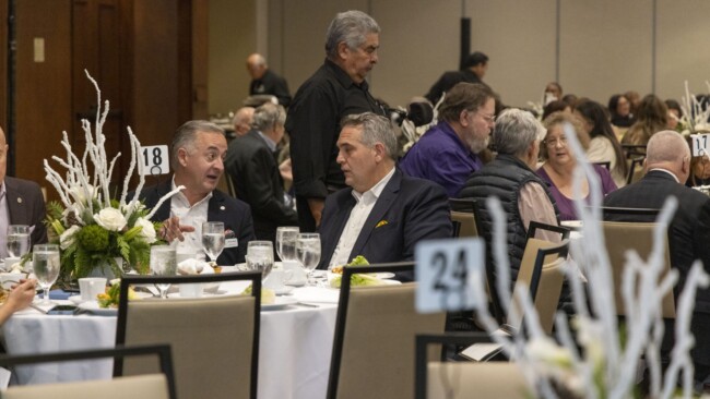 A large group of people sitting at tables during a formal event. Two men in suits are engaged in conversation at a table marked with the number 18. Floral centerpieces decorate the tables, and other attendees are visible in the background.