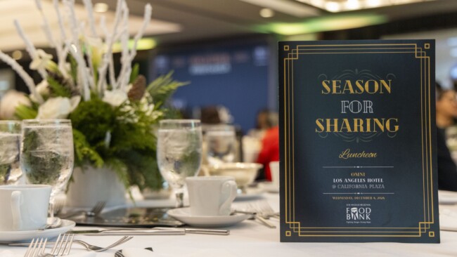 A decorated table at an event with a sign reading "Season for Sharing Luncheon" at the Omni Los Angeles Hotel. The table features floral arrangements, glassware, and cutlery. In the background, blurred attendees are visible.
