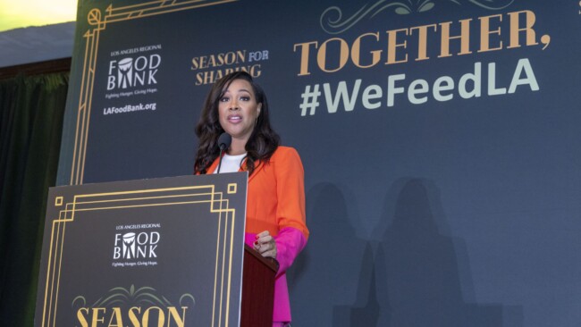 A woman in a colorful outfit speaks at a podium during a Los Angeles Regional Food Bank event. The backdrop displays text: "TOGETHER, #WeFeedLA" and "Season for Sharing.