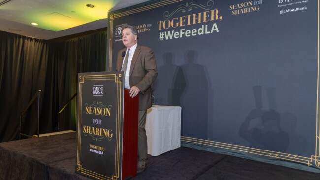 A man in a brown suit speaks at a podium with a backdrop reading "Together, Season for Sharing, #WeFeedLA" and "Food Bank." The stage is set with a classy design in a dimly lit room.