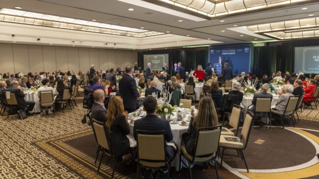 A large group of people is seated around round tables in a conference room. The room is decorated with white tablecloths and floral centerpieces. A stage with a podium and presentation screens is at the front. Attendees are dressed in formal attire.
