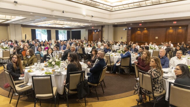 A large audience sits at round tables during a formal event in a spacious banquet hall. The tables are set with white tablecloths, plates of food, and floral centerpieces. Attendees are dressed in business attire, focused on the front of the room.
