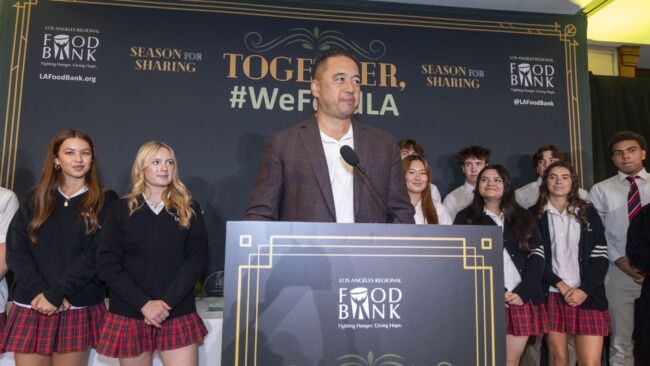A man stands at a podium labeled "Food Bank" addressing an audience. Behind him are a group of young people in school uniforms. The backdrop reads "Together, #WeFeedLA" and "Season for Sharing.