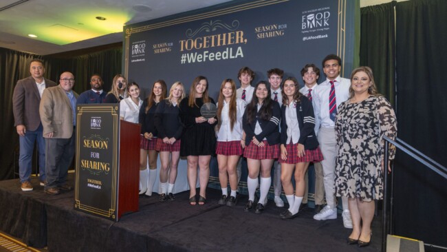 A group of students in matching uniforms stands on stage with several adults at a "We Feed LA" event. They are holding an award. The backdrop is themed with Season of Sharing logos and a hashtag: #WeFeedLA.
