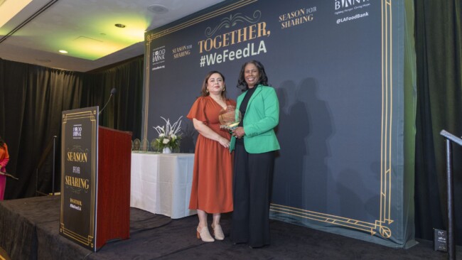 Two people stand on a stage, one holding a trophy. They are posed in front of a backdrop with the text “Together, #WeFeedLA.” The event appears formal, with flowers and a podium visible in the background.