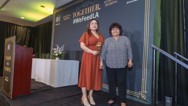 Two women stand on a stage, one holding a trophy. They're in front of a backdrop with the text, "Together, #WeFeedLA." A decorated table is in the background. The setting appears to be a formal event or ceremony.