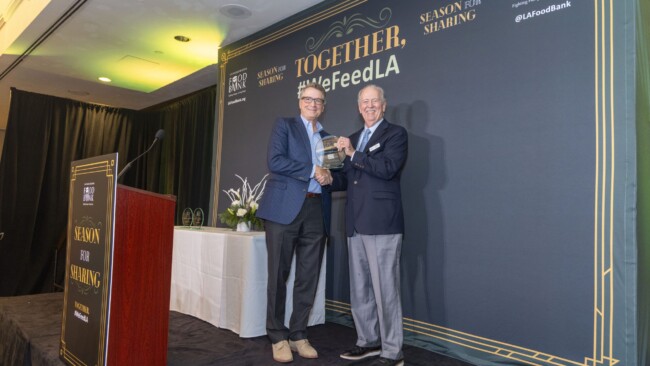 Two men in suits shake hands on a stage at the #WeFeedLA event. One holds an award, smiling. The backdrop displays the event theme, "Together, A Season for Sharing," and floral arrangements decorate the stage.