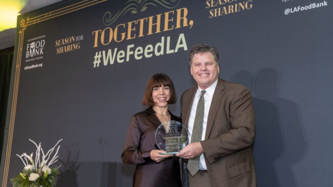 A man and woman are standing together at an event, holding an award. The backdrop displays the text "Together, #WeFeedLA" and logos for the LA Food Bank and Season for Sharing. There is a small floral arrangement to the side.