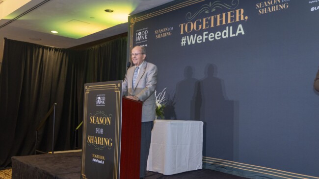 A man in a suit stands at a podium with the text "Season for Sharing" in front of a decorative backdrop reading "Together, #WeFeedLA." There is a floral arrangement on a small table beside the podium.