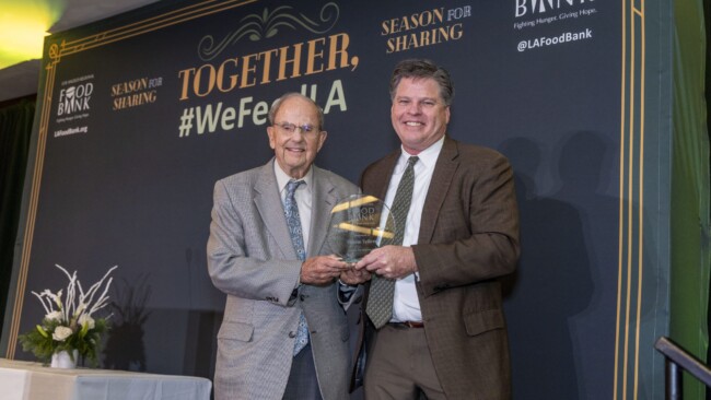 Two men smiling and holding a glass award in front of a backdrop with text: "TOGETHER, #WeFeedLA, Season for Sharing, LAFoodBank." A decorative plant is on a table to the left.