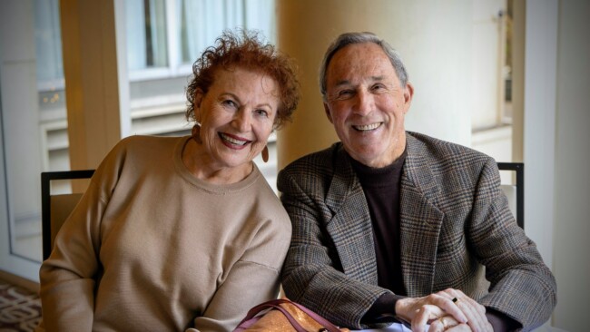 An elderly couple smiles warmly at the camera while sitting indoors, embodying the spirit of community supported by the Los Angeles Regional Food Bank. The woman wears a beige top and hoop earrings, while the man dons a checked blazer with a dark turtleneck. A window with blinds is seen in the background.