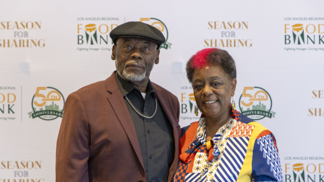 A man and woman are standing together, smiling in front of a "Los Angeles Regional Food Bank Season for Sharing 50th Anniversary" backdrop. The man sports a hat and blazer, while the woman dazzles in a colorful patterned outfit, embodying the festive spirit of the event.