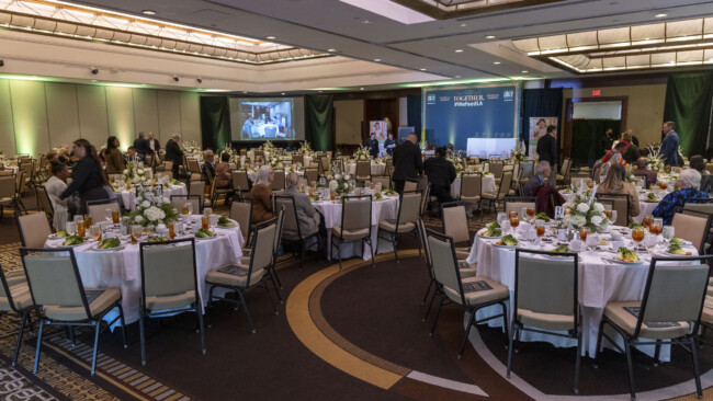 A large banquet hall set for a formal event hosted by the Los Angeles Regional Food Bank. Round tables with white tablecloths boast floral centerpieces, tableware, and glasses. Guests are seated and mingling as a stage and screen loom in the background.