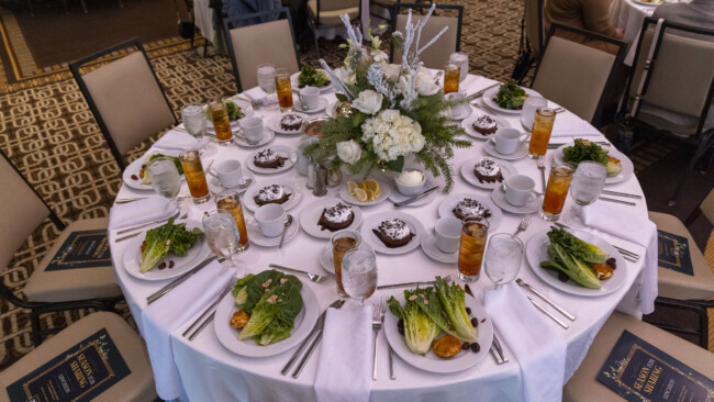 A round dining table set with white linens, elegantly adorned with a floral centerpiece, evokes the spirit of the Los Angeles Regional Food Bank. The table boasts plates with salads, dessert dishes, iced tea, water, and cutlery. Each seat has a menu placed on the chair.