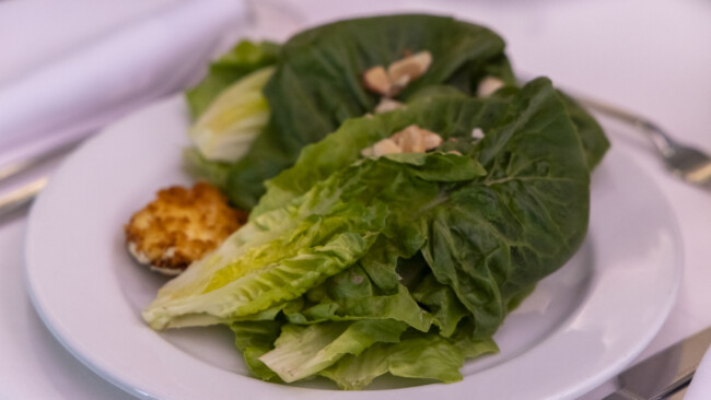 A white plate with a fresh green salad made of lettuce leaves, topped with almond slices, and accompanied by a small pastry on the side supports the efforts of the Los Angeles Regional Food Bank in promoting nutritious meals for everyone.