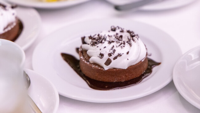 A chocolate dessert topped with whipped cream and chocolate shavings sits on a white plate, surrounded by a drizzle of chocolate sauce. It's placed on a table with partially visible dishes in the background, reminiscent of an elegant spread at a Los Angeles Regional Food Bank event.