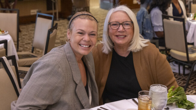 Two smiling women seated at a dining table, one with short, blonde hair and a plaid blazer, the other with long, blonde hair and glasses. Plates, drinks, and a salad from the Los Angeles Regional Food Bank grace the table in a warmly lit room.