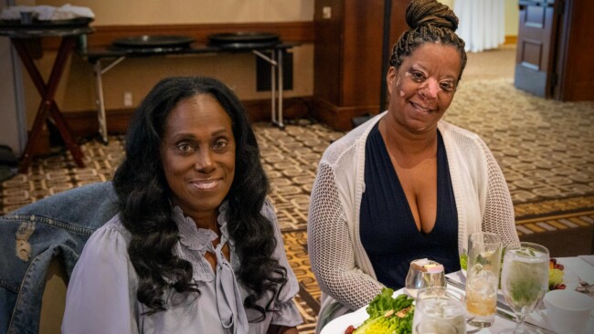 Two women are sitting at a table in a restaurant, enjoying a meal that supports the Los Angeles Regional Food Bank. The table is adorned with plates of food, drinks, and a colorful dish. One wears a light blouse while the other sports a white cardigan over a dark top, both smiling warmly at the camera.