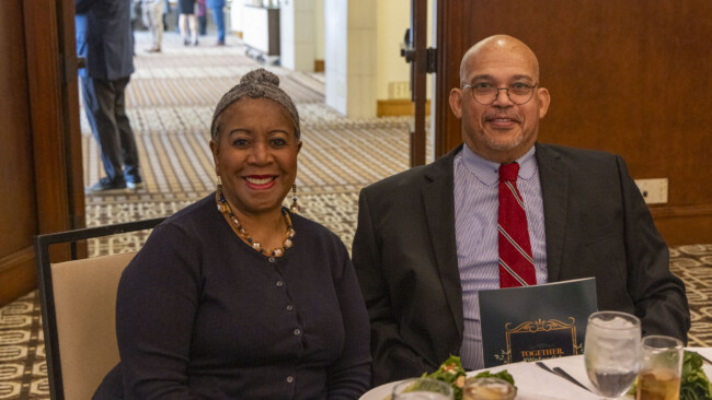 A man in a suit and tie and a woman in a black top sit at a table with plates of food and drinks, smiling at the camera. The warmly lit dining area, featuring patterned carpet, is part of an event supporting the Los Angeles Regional Food Bank.