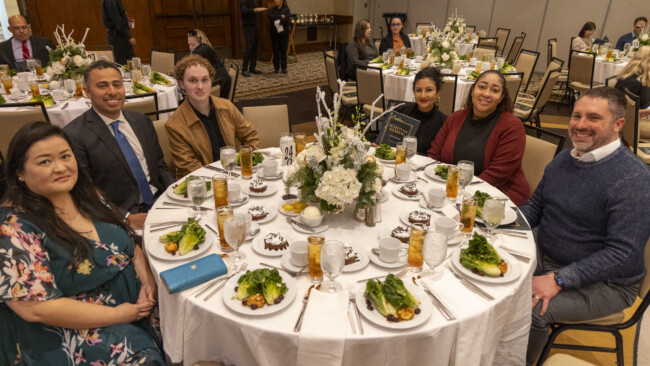 A group of people sit around a round banquet table set with salads, desserts, and drinks. The table features a floral centerpiece. Other guests and tables are visible in the background at this Los Angeles Regional Food Bank event.
