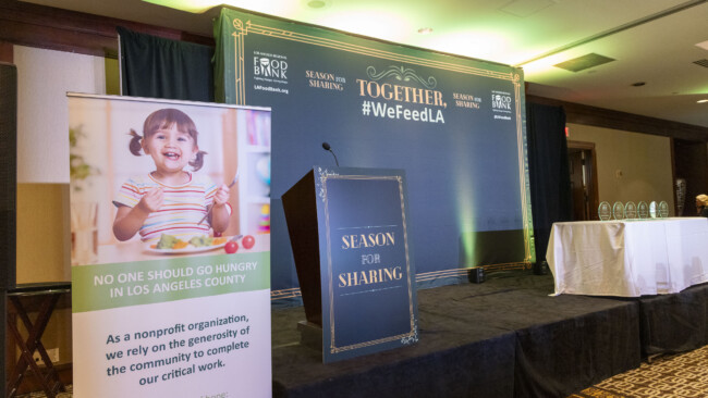 A conference room features a podium displaying "Season for Sharing." Nearby, a banner with a smiling child declares, "No one should go hungry in Los Angeles County." Awards are arranged on a table, while the backdrop proudly highlights "#WeFeedLA" and "Together" alongside the Los Angeles Regional Food Bank.