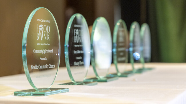 Several glass awards are lined up on a white table, each inscribed with "Food Bank Community Spirit Award" and reflecting light. The closest award honors "NewLife Community Church" for its efforts in collaboration with the Los Angeles Regional Food Bank.